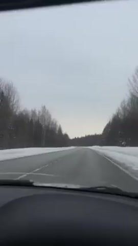 The video shows the view from inside a car driving on a straight, snowy road. There are forests on either side, also covered in snow. The sky is overcast and grey. The dashboard and parts of the car interior are visible in the foreground. There is music playing in the background.