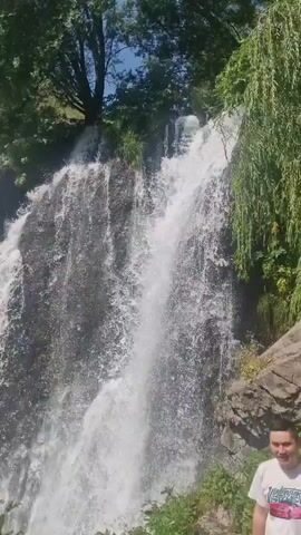 A man stands in front of a waterfall, smiling. The waterfall is surrounded by lush green trees and rocks, with a clear blue sky above. The water cascades down a rocky cliff face, creating a misty spray. The man is wearing a white t-shirt and appears to be enjoying the scenery.