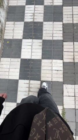 The video shows a first-person perspective of someone walking on a tiled pavement. The pavement is made up of alternating rows of grey and white tiles, laid out in a checkerboard pattern. The person is wearing dark denim jeans, white sneakers with dark trim, and a dark-colored garment. A brown Louis Vuitton patterned bag is visible on the right side of the frame, close to the person's hip. The person is walking at a moderate pace. Small amounts of dirt and vegetation can be seen along the edges of the pavement.