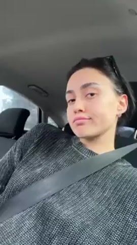 A young woman is sitting in the passenger seat of a car, wearing a gray sweater and a seatbelt. She has dark hair pulled back and is looking directly at the camera. She smiles and briefly holds up her fingers in a peace sign. The car interior is visible, including the black seats and the dashboard. There's music playing in the background.  The scene is well-lit, and the video appears to be shot with a mobile phone. Through the car window, one can see trees.