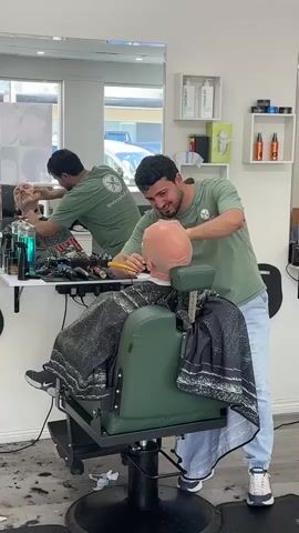 This video shows a barber shaving a client's head. The barber, wearing a green t-shirt and blue jeans, is using a straight razor to shave the client's head. The client, who is bald, is seated in a green barber chair and is wearing a black and white patterned cape. The barber is smiling and appears to be concentrating on his work. The barber shop is clean and well-lit, with shelves of grooming products in the background. A mirror reflects the scene, showing the barber and client from another angle. The floor is covered with hair clippings.