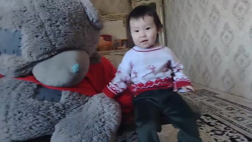 A young child sits on a stool next to a large gray teddy bear wearing a red shirt. The child is wearing a white sweater with a reindeer pattern and dark-colored pants. The room has wallpaper with a repeating pattern and a rug with a geometric design. In the background, there is a wooden cabinet with a mirror. The child raises their arm and smiles at the camera.
