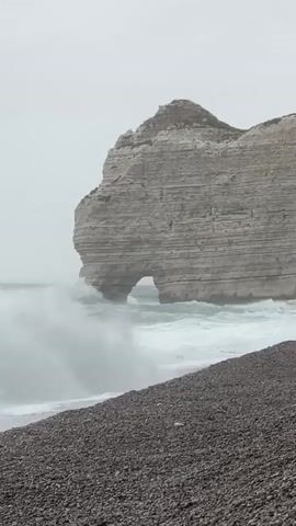 A drone shot shows waves crashing on a rocky beach near a large cliff with a hole in it.