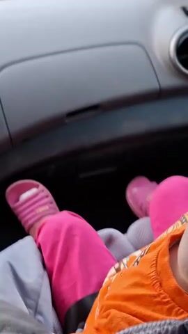 A close-up video shows a young child, likely a toddler, in the passenger seat of a car. The child has blonde hair pulled back with a pink hair tie, is wearing an orange t-shirt and pink leggings. The child's face is framed by the car's interior, with a gray dashboard and air vent visible in the background. The child's expression shifts from a slight smile to a more playful look, with their tongue sticking out slightly. The video appears to be shot from the perspective of someone sitting next to the child, possibly the driver or another passenger. The overall tone is casual and suggests a candid moment captured during a car ride.