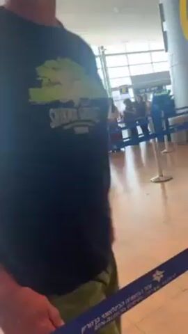 The video shows a man in an airport. He is wearing a black t-shirt with a green logo, green pants, and a pink hat. He stands in a queue lined with stanchions and blue straps. In the background there are people waiting in line with their suitcases. The video turns downwards towards the floor, and the man is shown in a crouched position. The video returns upwards to show the queue again.