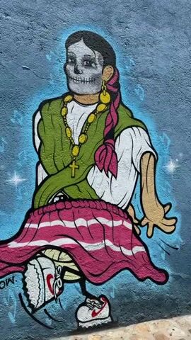 The video pans along a mural depicting Day of the Dead figures on a blue wall next to a cobblestone street. The mural features cartoonish skeletons playing musical instruments and a woman in traditional attire with a skull face. The figures are adorned with colorful details and surrounded by floral motifs. The wall has red window frames with metal bars. The video moves along the street, revealing more of the mural and the surrounding buildings.