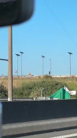 The video is a first-person perspective from inside a vehicle, likely a car, looking out the passenger window. The scene is a highway or a road with a clear blue sky. Several tall, thin lampposts with solar panels on top are visible along the roadside. There is a green, grassy embankment with some bushes. A diamond-shaped white sign is partially visible on the embankment. In the distance, a truck with "Ewals Cargo Care" written on its side is seen. An overpass with cars driving on it is also visible. The video continues to show the road and the surrounding landscape, with more lampposts and signage. A truck's cab with the Scania logo is briefly visible. The camera appears to be moving forward, indicating the vehicle is in motion.