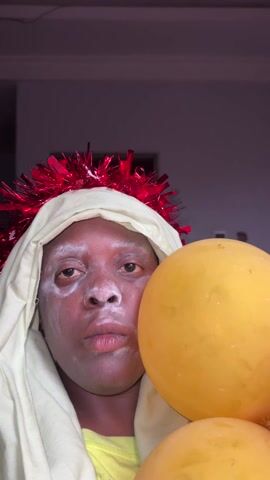 The video shows a person with dark skin wearing a white hooded shirt and a red tinsel garland on their head. The person's face has a white powdery substance on it. They are holding several colorful balloons. The background is a plain wall. The person looks directly at the camera with a neutral expression. The camera angle is slightly below eye level, looking up at the person. The lighting is somewhat dim, but sufficient to see the details.