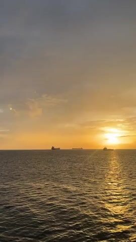 The video shows a sunset over the ocean. The sun is low on the horizon, casting a golden glow across the water. There are three ships visible in the distance, silhouetted against the bright sky. The sky is mostly a mix of orange and yellow near the horizon, with some darker grey clouds above. The ocean is calm with small ripples. The camera is relatively still.