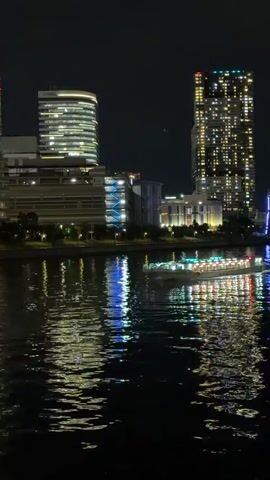 A video captured at night shows a cityscape with illuminated buildings and a body of water. A boat with numerous lights travels across the water, reflecting the lights of the city. The buildings are tall, with many windows displaying various colored lights. The water is dark, with shimmering reflections of the lights from the buildings and the boat. The overall scene depicts a vibrant urban environment at night.