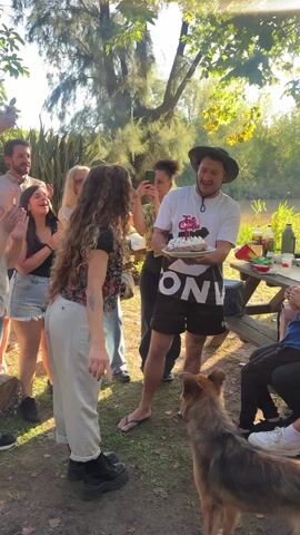 A birthday celebration unfolds outdoors, with a group of about a dozen people gathered around a picnic table. They are clapping and singing "Happy Birthday". A woman with long, curly hair and wearing a floral vest and white pants stands in front of a man holding a cake with candles, who’s also wearing a hat and a t-shirt.  A dog stands nearby, curious about the cake. Several people are filming the scene on their phones. The picnic table is covered with food and drinks. The setting appears to be a park or backyard, with trees and greenery in the background.