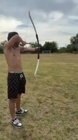 A man stands shirtless on a grassy field and shoots an arrow from a recurve bow toward a target. He is wearing black and white patterned shorts and matching sneakers. The target is green with a white, orange, and black bullseye. There are trees and soccer goals in the background under a cloudy sky. After the arrow is released, the man steps to the side, gesturing with his hands as if in explanation.