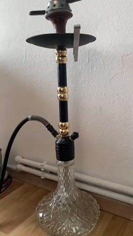 A hookah is shown. It is clear glass with a black and gold stem. A black hose is attached to the side. The top of the hookah has a dark brown bowl with a black lid and charcoal tongs resting on the side. Smoke emanates from the top of the hookah. The hookah is placed on a wooden floor in front of a white wall. There are white baseboard heaters behind the hookah.