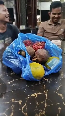 The video shows a close-up of a blue plastic bag filled with various fruits, including rambutan, mangosteen, and citrus fruits like oranges or lemons. The bag is placed on a dark, reflective table. In the background, two men are partially visible, seated at the table. The man on the left is laughing heartily, while the man on the right is smiling and looking towards the bag of fruit. The environment appears to be indoors, possibly a restaurant or a market, with blurred background elements suggesting a casual setting. The lighting is dim.