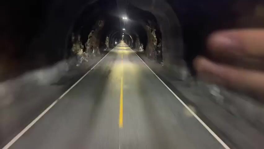 The video is a first-person perspective traveling through a dark tunnel. The road is asphalt with a yellow line down the center and white lines on the sides. Lights are visible intermittently along the ceiling of the tunnel, illuminating the rough, rocky walls. The tunnel appears to be long, with the light at the end becoming increasingly visible as the perspective moves forward. There are no other vehicles or individuals visible in the video.