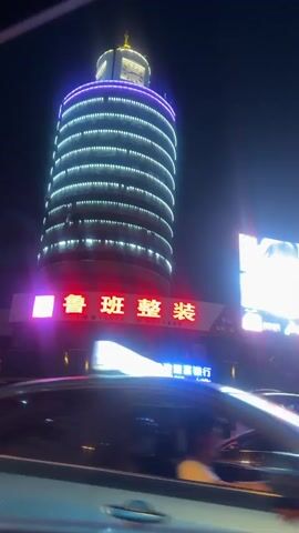 The video shows a night-time street scene in China. In the background, a tall, cylindrical building with horizontal bands of white lights stands out. Below the building are neon signs in Chinese and English, with the English sign indicating a Postal Savings Bank of China branch. Cars are parked along the street in front of the building, and vehicles move in the foreground, including the car from which the video is shot. A car horn sounds intermittently.  A billboard displaying "5G" is visible.