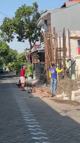 Three men are working on a construction site outdoors. Two men are standing and appear to be installing wooden posts, with one man in a blue and yellow shirt holding a post. The third man is kneeling and is near a pile of wood. The construction site has a pile of sand and bricks. The background shows a building with a white brick wall. A street with patterned pavement is in the foreground.
