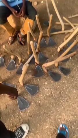 The media file shows a collection of hoes laid out on the ground. A young boy wearing a blue collared shirt and black pants is sitting on a rock amidst the hoes, appearing to be working on them or supervising them. The hoes are all made with metal blades attached to wooden handles. The setting appears to be outdoors, with sandy or dusty ground and a building in the background. There is also a motorcycle visible in the background. The lighting suggests it is daytime, possibly late afternoon given the shadows. The boy's expression is focused. The hoes are the primary objects in the scene, and their arrangement suggests they are either being made, repaired, or prepared for use. The location is identified as Reak, Nigeria. There is text visible on the boy's shirt, but it is too small to read.