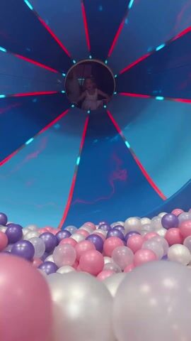 A young girl slides from a tunnel into a ball pit. She is wearing a white sleeveless dress. The ball pit is filled with pink, purple, and white balls. The tunnel is lined with blue fabric and red and light blue LED lights. The girl smiles as she lands in the ball pit. Another child is visible at the top of the tunnel.