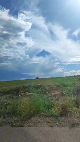 The video shows a view from a moving vehicle. The scene is a grassy hillside under a partly cloudy sky. A road is visible in the foreground. In the distance, a small structure or monument stands on the crest of the hill. As the vehicle moves, the perspective shifts to reveal more of the landscape, including a distant building or complex and a fence line. The overall environment appears to be rural or undeveloped.