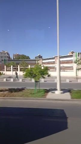 The video shows a view from a moving vehicle driving along a road on a sunny day. Buildings are visible on the opposite side of the road, with a strip of green grass and small trees separating the road from the sidewalk.  A person in a white robe walks along the sidewalk and another person in a blue shirt walks a little further ahead.  Several cars are parked in the parking lot next to the sidewalk. The sky is clear and blue.  The video captures a brief scene of urban life.