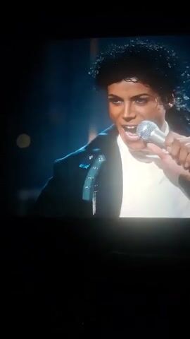 The video shows a man, likely Michael Jackson, performing on stage. He is wearing a white t-shirt, a black jacket with silver embellishments, and dark pants with a white stripe. He is holding a microphone and appears to be singing. Behind him, other musicians are visible, one playing a keyboard. The stage is lit with blue and white lights, creating a dramatic atmosphere. The background appears to be a grid-like structure with horizontal blue lights. The setting is a concert or live performance. There is no visible text in the video. The time of day and weather are not discernible. The emotion conveyed by the performer seems to be one of energetic engagement with the audience.