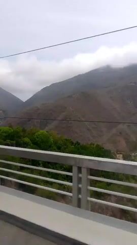 The video shows a view of mountains from a bridge. The mountains are mostly brown with patches of green trees. There is a bridge in the distance to the left. The sky is overcast with white clouds. The camera is moving, suggesting that the video was taken from a vehicle crossing the bridge. There is music playing in the background.