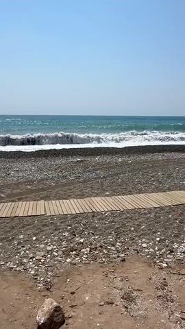 The video shows a beach scene. In the foreground, there is a wooden pathway made of planks. The ground consists of sand and small pebbles. Further back, a strip of dark pebbles meets the sea. The sea itself is a vibrant blue-green with waves breaking onto the shore, creating white foam. A red flag is visible on a pole planted in the pebble area near the water's edge. In the distance, the horizon is a clear blue sky, with a hint of a coastline featuring some buildings and palm trees on the left side of the frame. The overall atmosphere is one of a sunny day at the beach.