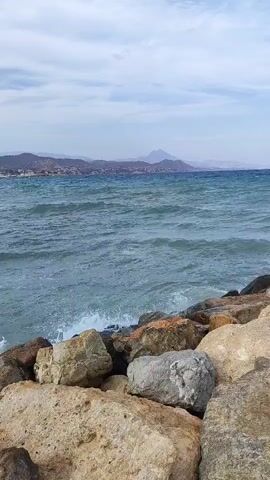 The video shows a rocky shoreline with the ocean in the background. The water is a turquoise blue and there are waves crashing against the rocks. In the distance, there is a coastline with hills and buildings. The sky is cloudy.