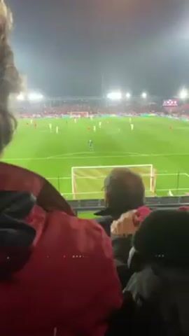 The video shows a football game being played at night from the perspective of a spectator in the stands. The spectator is sitting near other people, as seen by a head and a red jacket in the foreground. The field is well-lit by stadium lights, and the players are wearing red and white uniforms. There are a significant number of fans in the stands, but their faces are not clearly visible due to the distance and lighting. The camera is mostly still, but there are slight movements due to the person holding the camera. The overall setting is that of an active sporting event with a lively atmosphere.