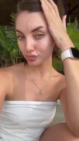 A woman in a strapless white top is outdoors. She is fair skinned with brown hair pulled back in a bun and wears a silver necklace and a white smartwatch. She is looking into the camera and touches her hair. The background is out of focus but shows trees, umbrellas, and people. The video appears to have been taken in a tropical area. A song with a dance beat is playing.