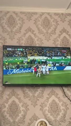 A television screen displays a soccer match. The score is 1-1. The time is 60:05. The teams are labeled BRA and COL. A group of players in white jerseys huddles on the field. The word "Michelob" is visible on a banner behind them. Another player in a green jersey with yellow stripes is shown in a close-up. He has a distinctive haircut. The background on the screen shows a crowd of spectators. The television is mounted on a wall covered with patterned wallpaper.