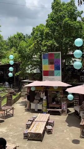 The video pans across an outdoor market or fairground. The scene is filled with stalls selling various items, likely crafts or art, given the banner visible in the background that reads "Living Art & Craft Series 2023". The stalls are adorned with pink umbrellas providing shade, and hanging from strings above are light blue paper lanterns, contributing to a festive atmosphere. Several tables, primarily made of bamboo, are set up in an open area, some with chairs or stools, suggesting a place for visitors to sit and relax. In the background, lush green trees and foliage create a natural backdrop, indicating the market is situated in a verdant environment. A few individuals can be seen at the stalls and in the surrounding area, though they are not the primary focus. The overall impression is of a lively, artistic, and community-oriented event taking place in an outdoor setting.