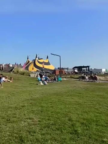 This is a short video clip showing a daytime outdoor scene under a clear blue sky. In the background, a large, striped yellow and black tent, possibly a circus tent, is prominent. To the right of the tent, there's a stage-like structure with some dark buildings behind it. In the middle ground, several people are seated on chairs and benches on a grassy slope, appearing relaxed and possibly watching something. Closer to the foreground, more people are spread out on the grass, some lying down. The environment is a grassy park-like area with gentle slopes. There is a black pole with a light fixture visible in the middle ground.