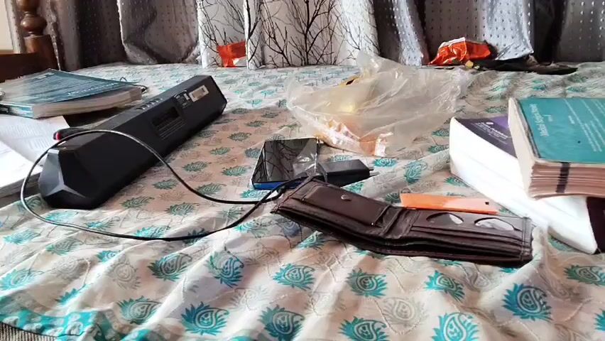 The video shows a cluttered bed with various objects scattered across it. In the foreground, a brown leather wallet is open, revealing coins and an orange object, possibly a small card or piece of plastic. To the left of the wallet is a black electronic device with a cord extending from it. Behind the wallet, a smartphone lies on the bed. Further back, a clear plastic bag contains what appears to be food items, and a stack of books is visible on the right side of the frame. A patterned bedsheet covers the surface. Curtains hang in the background. The lighting suggests an indoor setting, likely a bedroom during the daytime. There are no people visible in the video.