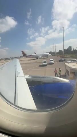 The video opens with a view from inside an airplane window, showing a tarmac scene. Two individuals in reflective vests stand on the tarmac, facing away from the camera, near a small building. Several vehicles, including white vans and cars, are parked on the tarmac. Another airplane, branded with "ARKIA" and featuring a distinctive tail design, is visible in the background. The sky is blue with scattered white clouds. The camera briefly pans to the right, showing the interior of the airplane's cabin, focusing on a wall and a portion of a seat. The scene suggests the initial stages of an airplane's journey, possibly boarding or preparing for departure, under a clear sky.