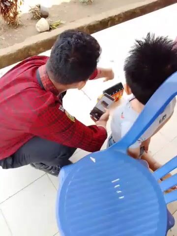 A young person wearing a red checkered shirt and dark pants kneels on a tiled floor, holding a smartphone. The smartphone screen displays a game or application. Another young person, wearing a light-colored shirt, stands next to the first person, looking at the smartphone. A blue plastic chair is partially visible in the foreground. The background shows a raised concrete planter box with some foliage. The scene appears to be outdoors, possibly a patio or walkway.
