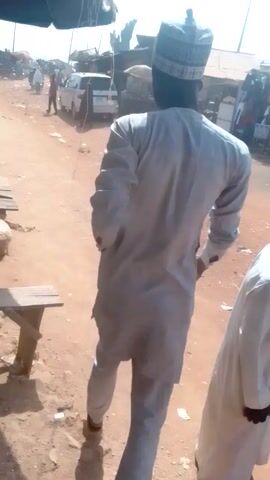 A person wearing a white robe and a patterned cap walks along a dusty road. The road is littered with trash. The person is holding a small brown object in their right hand.  A wooden structure is on the left side of the frame. In the background, there are other people and vehicles. The video is shot from a high angle, looking down.