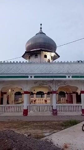 The video shows an exterior view of a mosque with a dome and arched walkways. The sky is overcast. There is a pile of dirt or rubble in the foreground. A person is visible in one of the arched walkways.