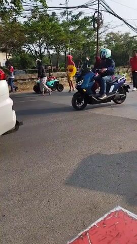 The video captures a street scene with several people and vehicles. In the foreground, a black and white motorcycle is parked or moving slowly on the right side of the road. Further down the road, a blue and white scooter carries two passengers, a woman in front and a child behind her. To the left of the scooter, a person in a red shirt and black pants walks with a red object, possibly a bat, in their hand. Behind them, a person in a yellow outfit and a red head covering walks. To the far left, a person on a dark motorcycle with a blue helmet rides past. Other individuals and a white object, possibly another vehicle, are visible in the background. The environment appears to be an urban or suburban area with trees lining the road and power lines overhead. The lighting suggests it is daytime.