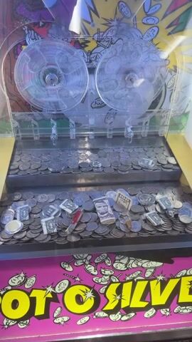 This is a close-up, overhead shot of a coin pusher arcade game. The game is filled with coins and some paper money. There are two rotating disks at the top of the machine, which are meant to push the coins forward. The machine has a yellow and pink color scheme, and the words "PUSH TO WIN" are visible on the front. The lighting is bright, and the scene appears to be indoors. There are no people visible in the video.