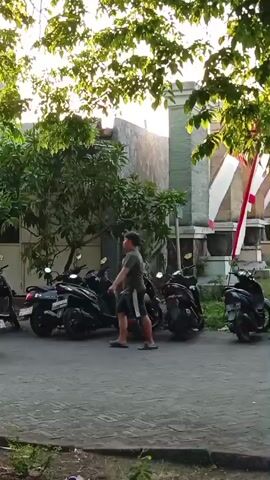 A group of motorcycles are parked in front of buildings decorated with red and white flags. Two people on a green scooter ride past, and another person is seen walking near the parked motorcycles. The environment appears to be an outdoor area with trees, a paved walkway, and a grassy area. The lighting suggests it is daytime, possibly morning or late afternoon.