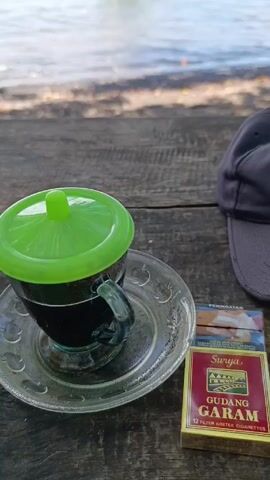 The video shows a view from a wooden table on a beach. On the table, there is a cup of dark liquid covered with a green lid, a pack of "Surya Gudang Garam" cigarettes, and a gray baseball cap. The camera pans up to reveal a turquoise sea with a small white boat anchored near the shore. A tree with green foliage hangs over the scene, partly obscuring the view. The sky is blue with some clouds. The beach appears to be sandy with some dark rocks or debris.