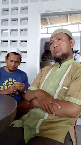 Several men are sitting around a round table. Some of them wear Muslim caps. There are glasses and cups on the table. The men are conversing. The room has a white wall with square openings for ventilation and a clock on the wall. A plant sits near a box labeled "Lauta."