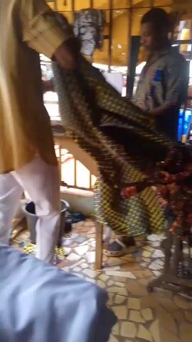 The video depicts a scene inside what appears to be a tailor's shop. Two men are visible, one wearing a striped shirt and white trousers, and the other wearing a patterned shirt and brown trousers. They seem to be working with pieces of fabric. A third person, wearing a pink shirt and red skirt, enters and exits the frame. The shop has a tiled floor and wooden furniture. A sewing machine covered with light blue cloth is in the foreground. Outside, the street is visible with vehicles passing by.