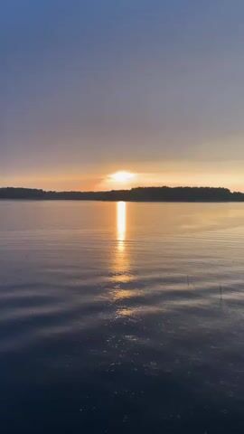 The video shows a beautiful sunrise or sunset over a lake with a line of trees on the horizon. The sun is bright, casting a golden reflection on the water's surface. The sky is a mix of dark blue and orange hues, suggesting either early morning or late evening. The water is relatively calm, with gentle ripples. There are a few small objects sticking out of the water near the lower right corner. Some indistinct speech can be heard.