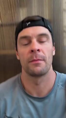 The video shows a man with short brown hair, wearing a black baseball cap and a grey long-sleeved shirt. He is looking at the camera and speaking. In the first few seconds, he touches his chin. He then smiles broadly at the camera before continuing to speak. The background appears to be a textured wall.