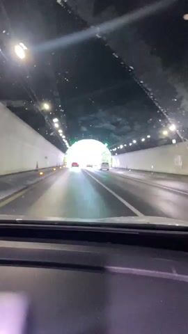 The video begins from the perspective of a car interior, looking out through the windshield. The car is driving through a tunnel, indicated by the artificial lighting on the ceiling and the curved walls. There are orange traffic cones visible on the side of the road. As the car moves forward, the tunnel opens up into daylight, revealing a two-lane road with green trees and vegetation on either side. The road appears wet, suggesting it has been raining or recently rained. More cars are visible on the road ahead. The sky is overcast and hazy, with mountains visible in the distance, partially obscured by fog or clouds. The video captures the transition from the darkness of the tunnel to the open, albeit gloomy, outdoor environment.