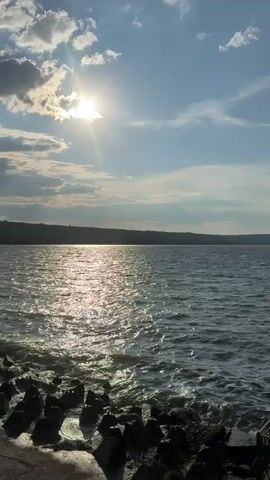 A vertical video shows a sunlit view of a lake or large body of water. The sun is bright and positioned in the upper left quadrant of the frame, partially obscured by clouds, casting a shimmering reflection on the water's surface. The water is choppy with small waves breaking against a rocky shore and a concrete embankment in the foreground. The opposite shore is lined with trees. The sky is blue with scattered white clouds. The video pans slowly from right to left.