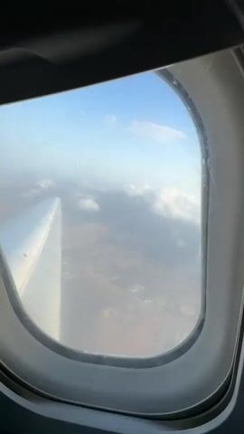 The video shows a view from an airplane window. Initially, the wing of the airplane is visible along with the ground below, appearing as a reddish-brown landscape. The view then shifts to focus primarily on thick, white clouds filling the frame. The sky above the clouds is a pale blue. The airplane window itself is slightly scratched and dirty.